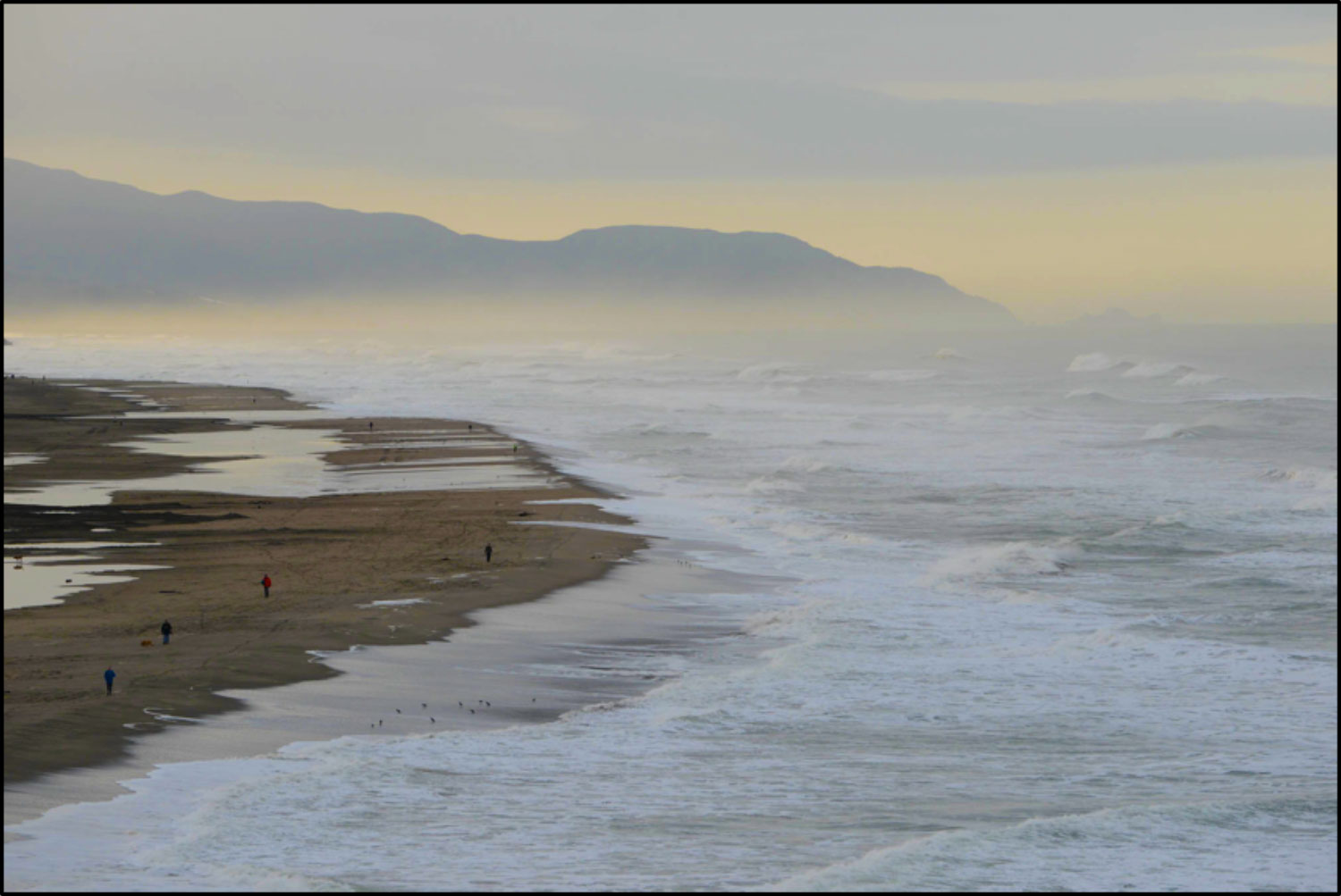 photograph of a coastline with waves, and people walking on the beach.